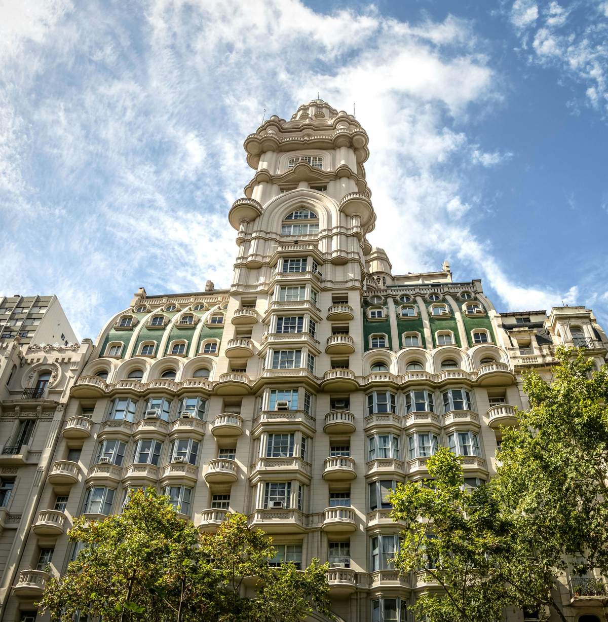 Palacio Barolo: Guided Tour Tall, ornate building with arched windows and decorative details, set against a blue sky with clouds and surrounded by trees.