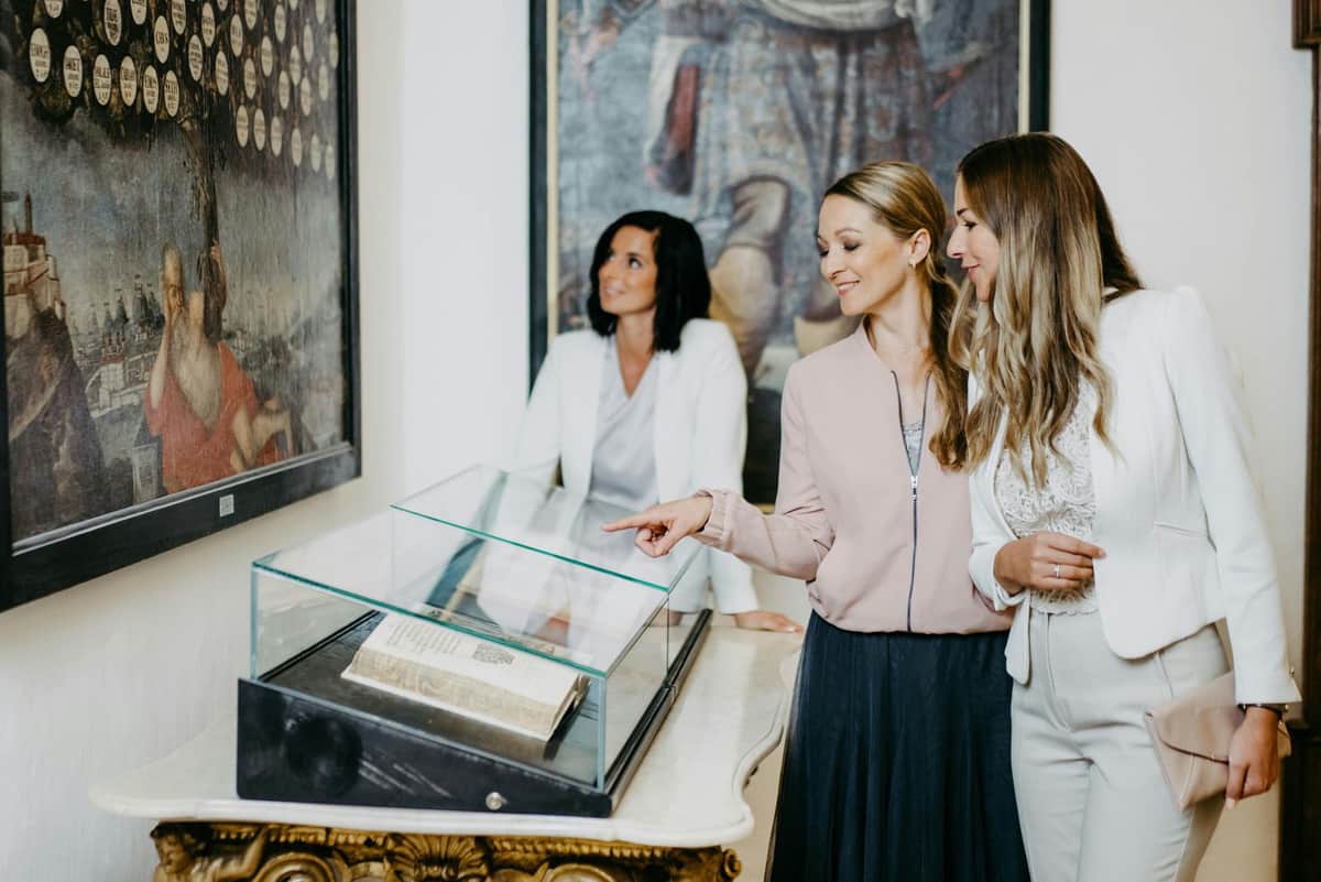 Forchtenstein Castle: Entry Ticket + Guided Tour Three women in smart attire admire a book displayed in a glass case in front of a painting.