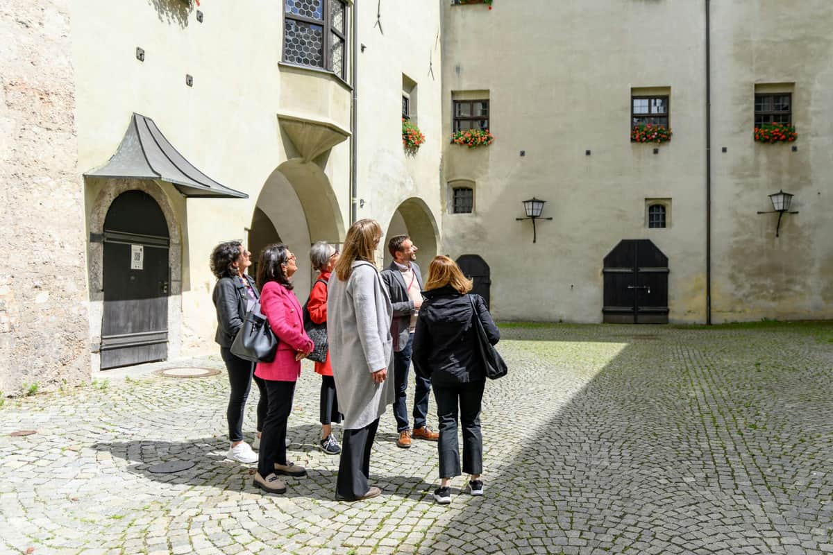A group of people standing in a cobblestone courtyard surrounded by historic buildings with arched doorways and flower boxes.
