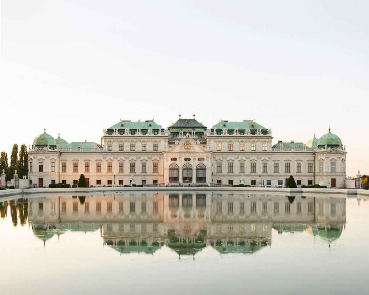 A large, ornate palace with a green roof is reflected in a calm body of water under a clear sky.
