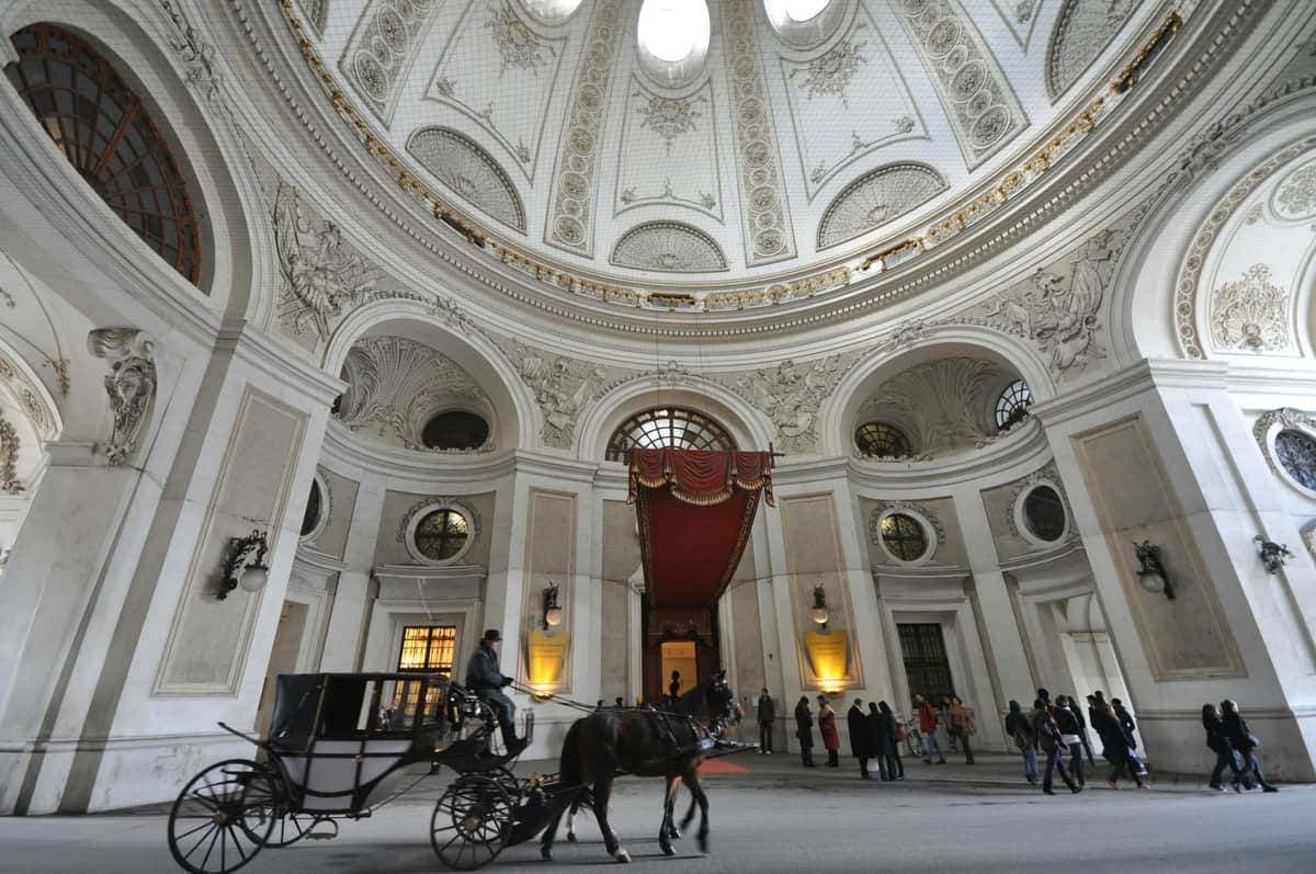 Palacio de Hofburg y Museo de Sisi: Tour guiado Un coche de caballos pasa bajo el gran techo ornamentado de un edificio abovedado con gente de pie cerca de la entrada.