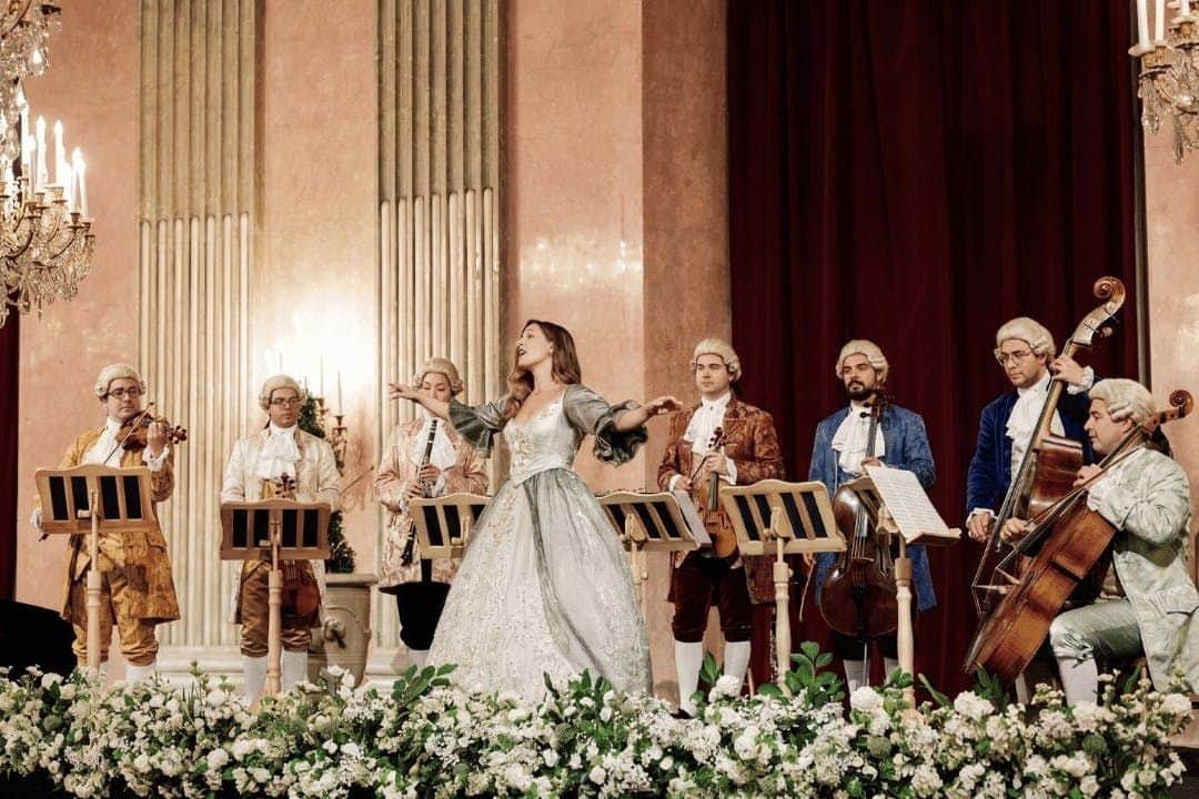 Palais Auersperg: Imperial Gala Concert A baroque-style ensemble with period costumes performs on stage. A soprano sings in front of musicians playing string instruments.