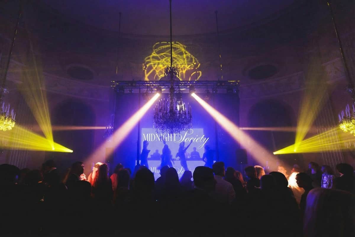 A dimly lit event with people gathered, stage lights, a chandelier, and a screen displaying 'Midnight Society.'