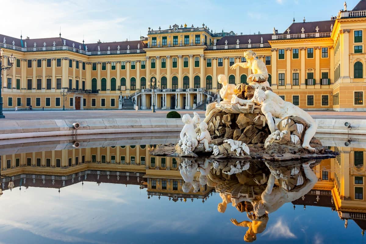 Ornate yellow palace with green windows reflected in a calm pool featuring a detailed white statue fountain in the foreground.