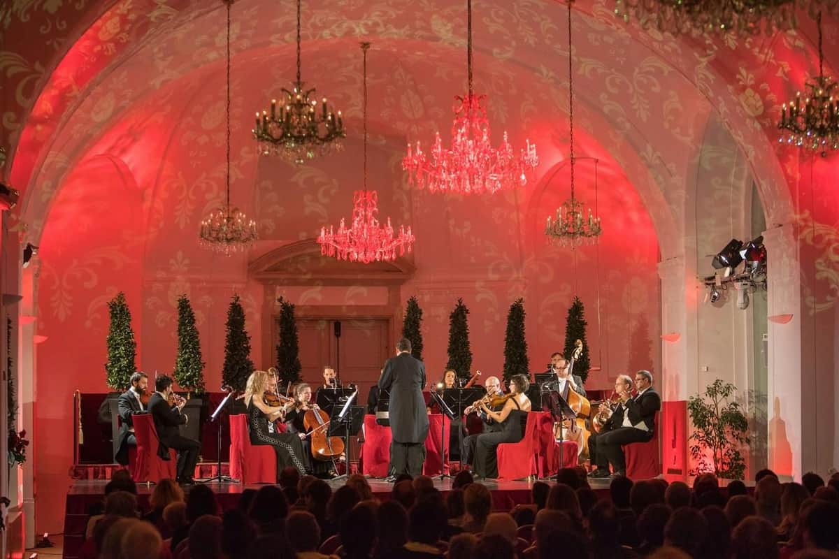 Palácio de Schönbrunn: Bilhete de Concerto Uma orquestra de câmara actua num palco com iluminação vermelha, candelabros e um cenário decorativo, vista por um público.