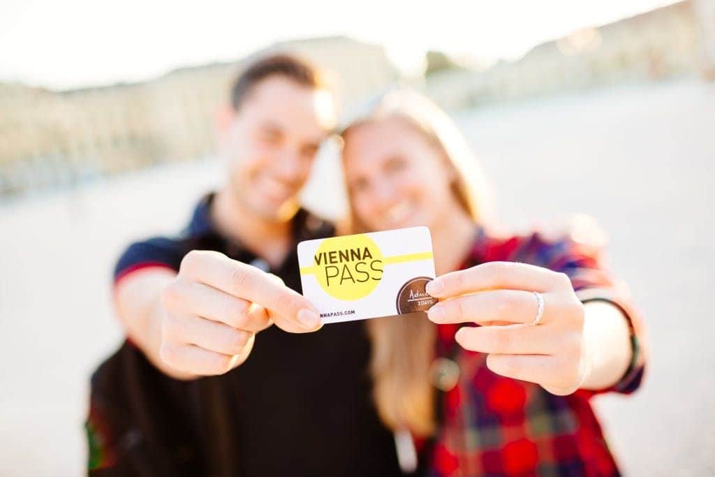 Two smiling people holding a Vienna Pass card, with blurred background indicating an outdoor setting.