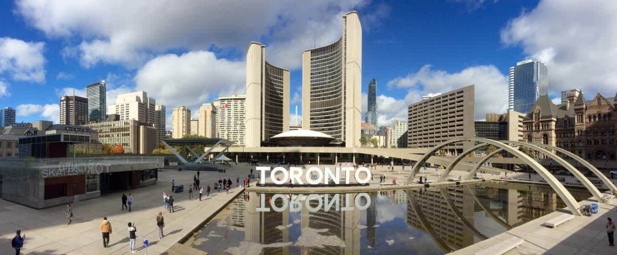 People walk around Toronto's City Hall with a large "TORONTO" sign and reflecting pool in the foreground.