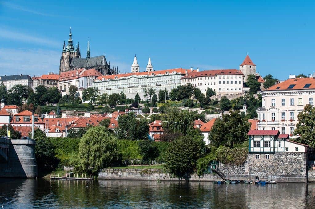 Panoramic view of historic buildings with red roofs, including a large cathedral, situated on a hillside above a river.