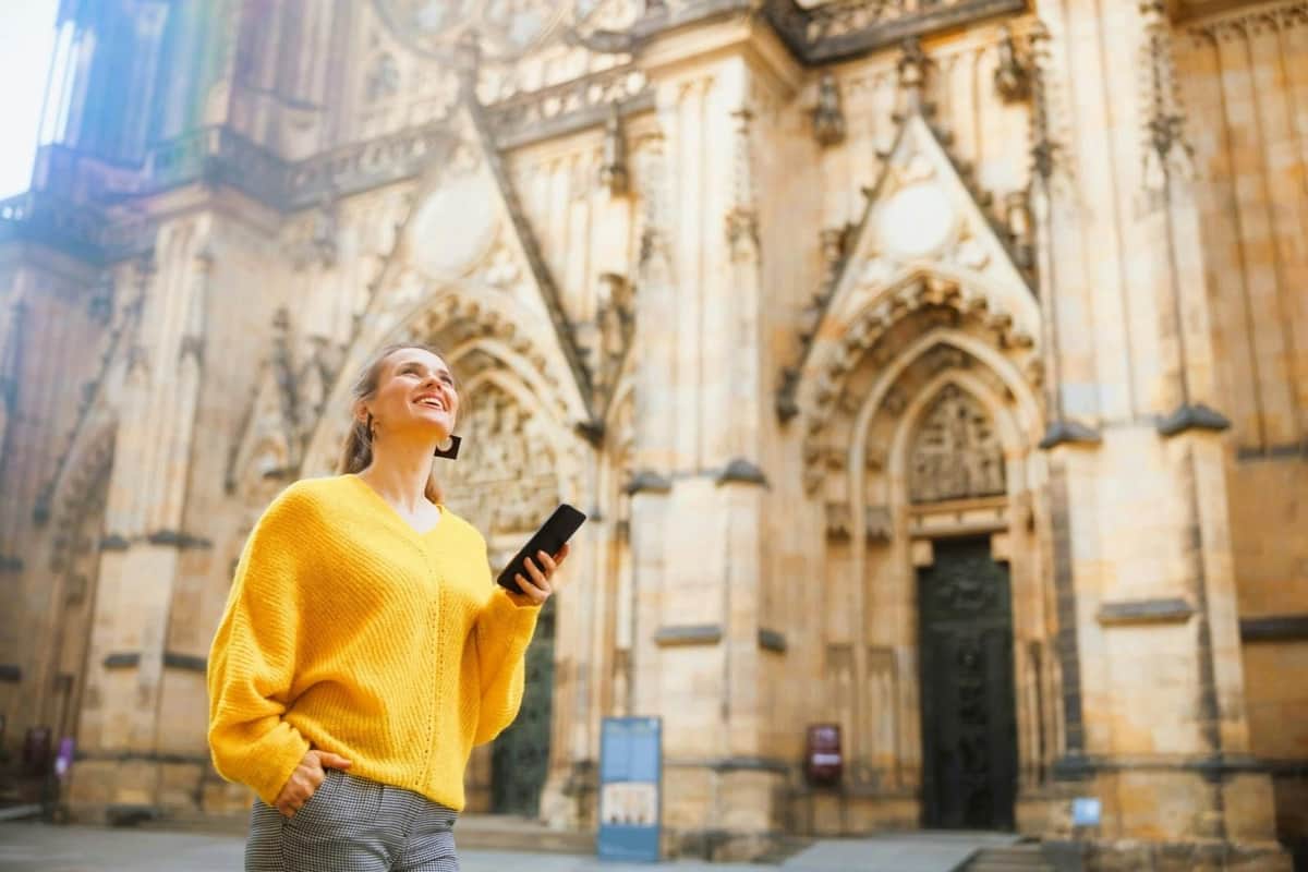A person in a yellow sweater holding a phone smiles while standing in front of an ornate historical building.