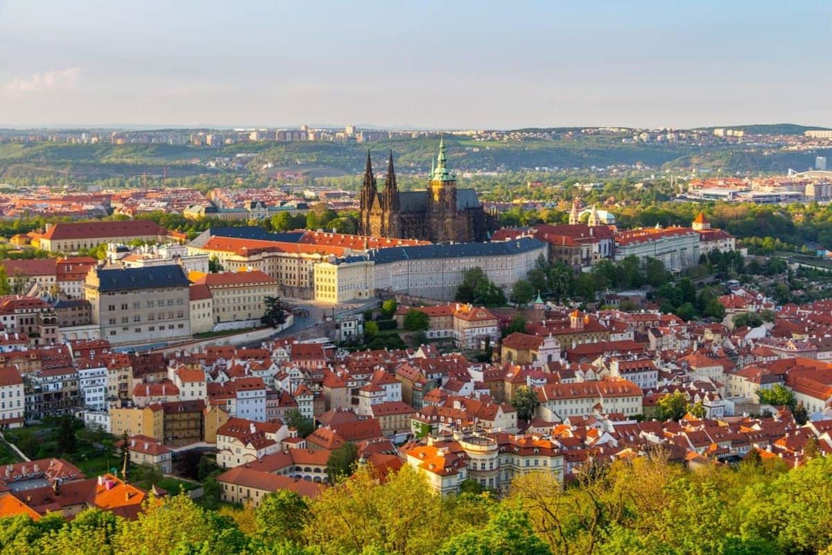 A panoramic view of a city with red-roofed buildings, a cathedral, and greenery under a clear sky.