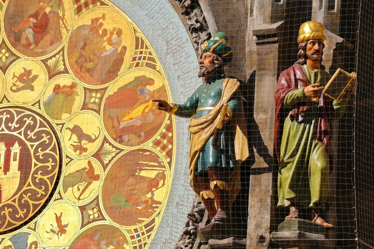 Prague Castle, Jewish Quarter & Astronomical Clock: Skip The Line + Audio Guide Statues of two figures in colorful attire holding objects, set against a medieval astronomical clock and stone facade.