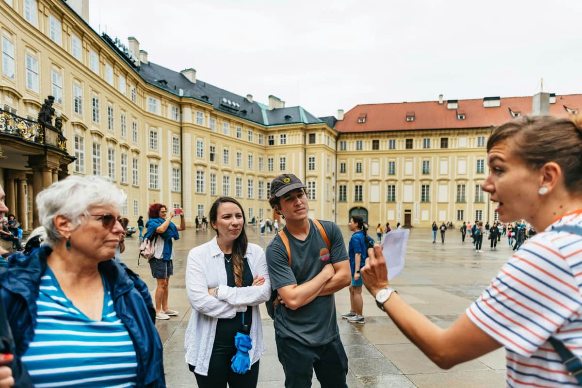 Prague: Guided City Bus Tour + Prague Castle Exterior Visit A group of people stands in a courtyard surrounded by historic buildings, one person appears to be leading or explaining something.