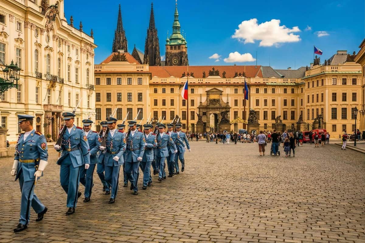 A procession of uniformed guards marching in a historic square with ornate buildings and a cathedral in the background.