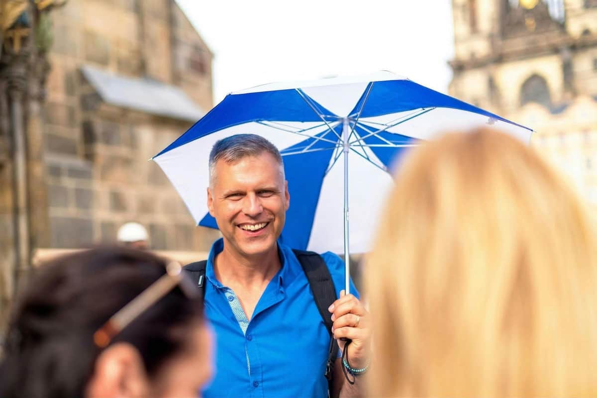 Um homem sorridente, de camisa azul, segura um guarda-chuva azul e branco ao ar livre. Em primeiro plano, vêem-se figuras desfocadas.