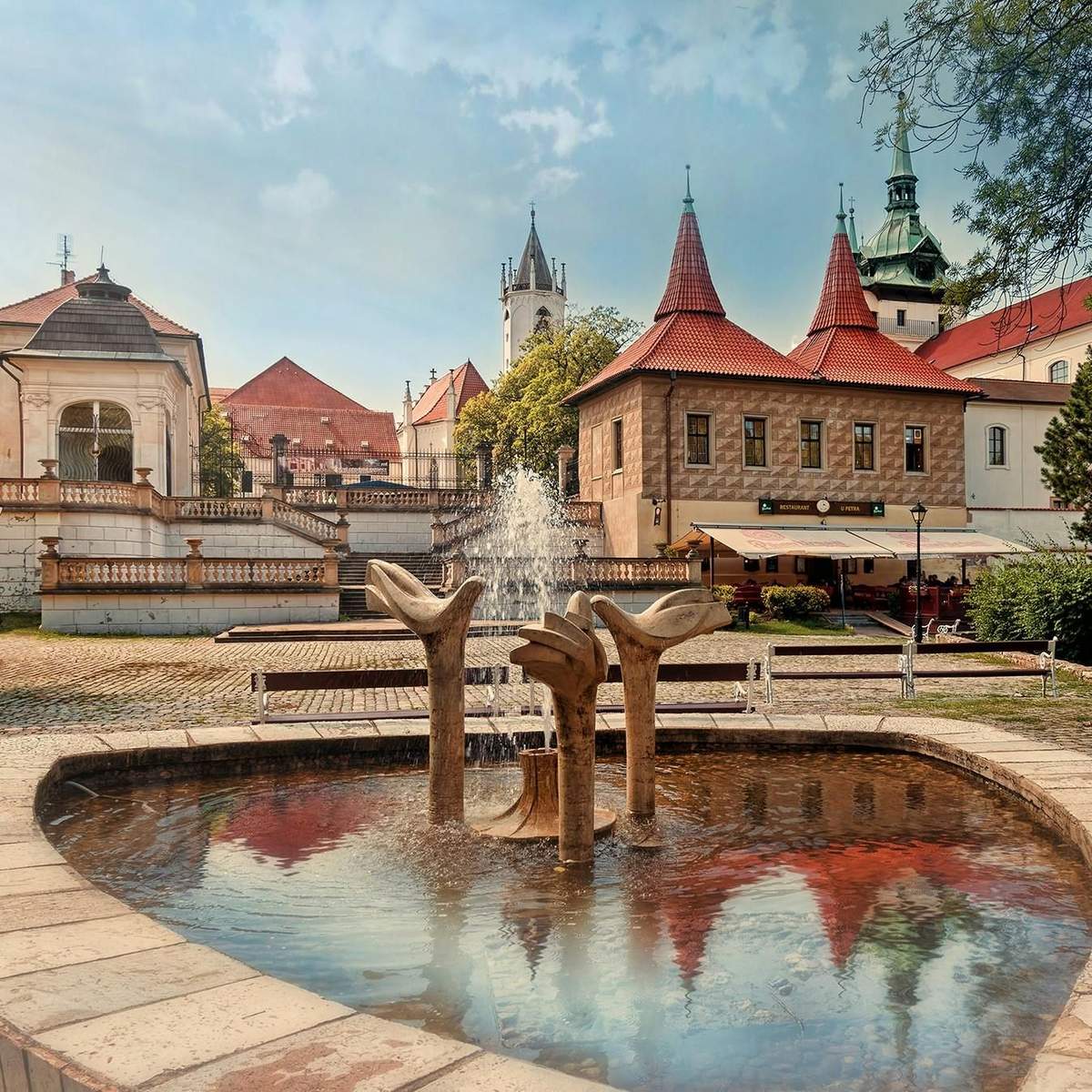 A stone fountain with water jets in a courtyard, surrounded by historic buildings with red roofs and spires.