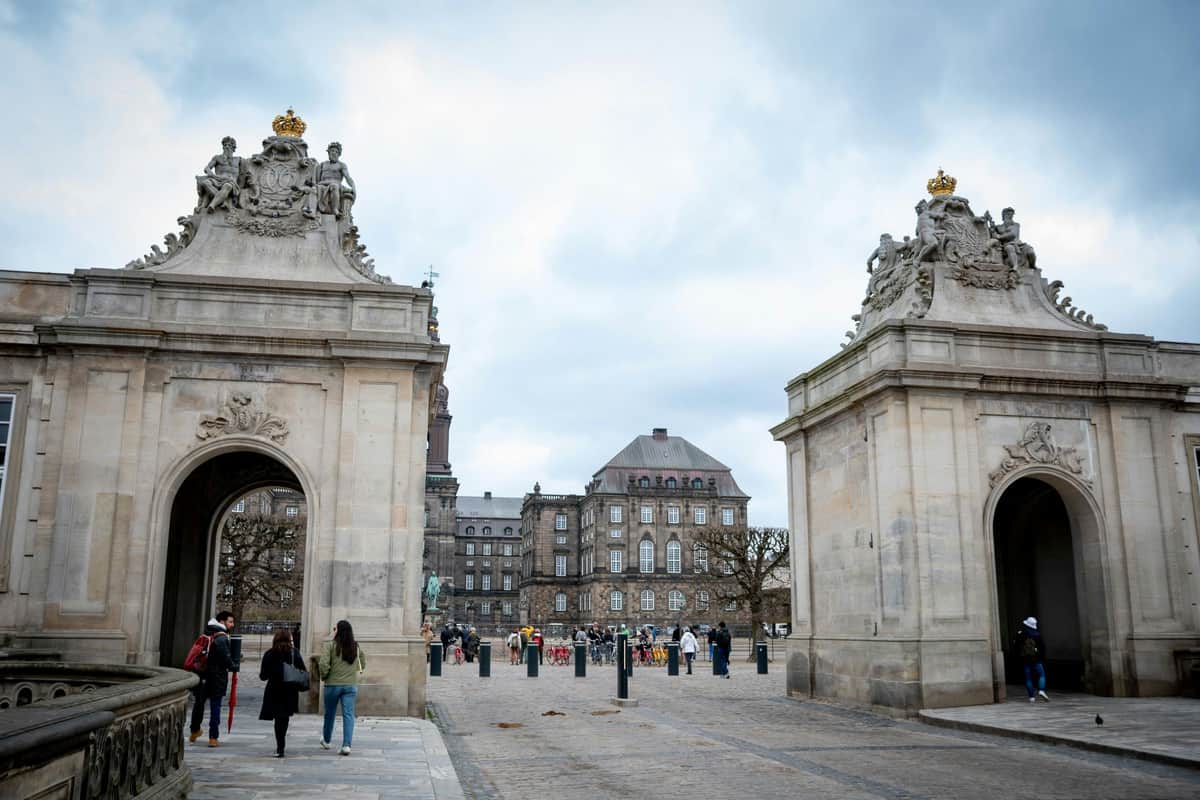 People walking between two ornate stone archways leading to a historic building under a cloudy sky.