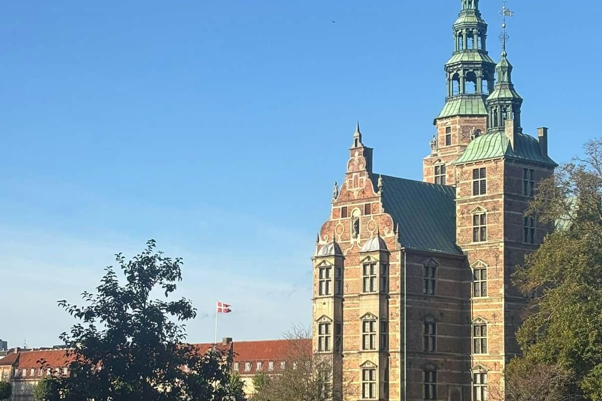 Historic castle with ornate towers and a red brick facade under a clear blue sky; a Danish flag flies in the background.