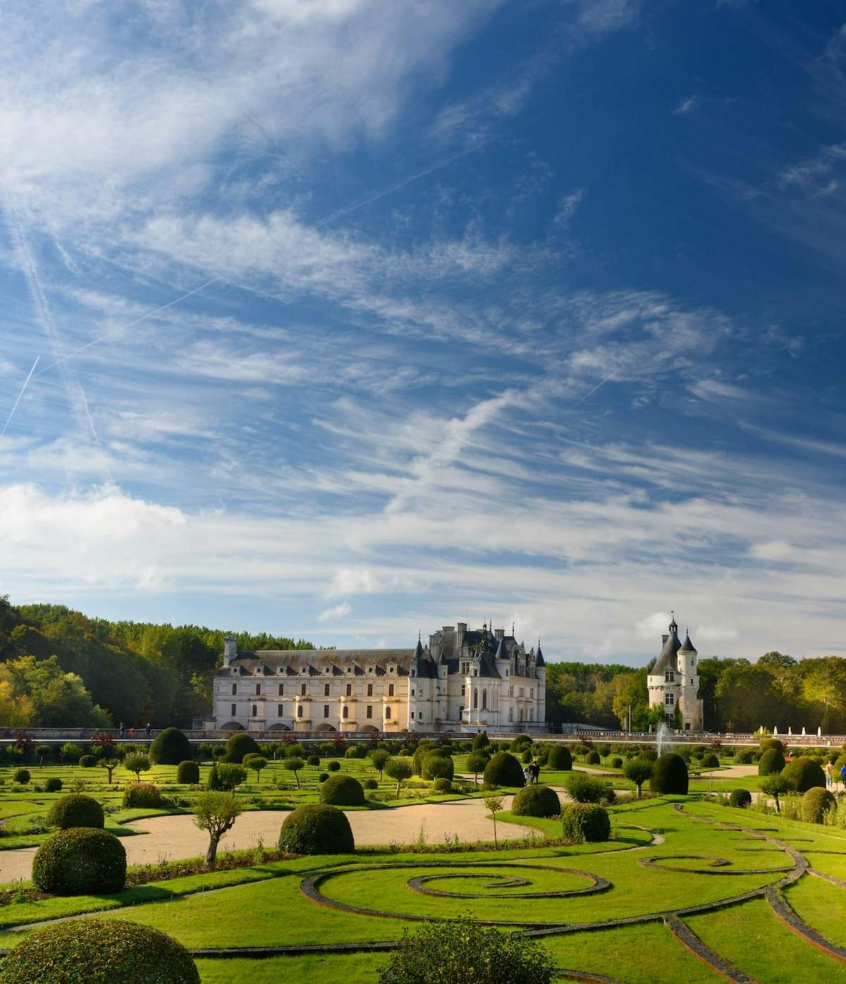 Château d’Amboise + Château du Clos Lucé + Château de Chenonceau A picturesque castle with landscaped gardens under a blue sky with scattered clouds and trees in the background.