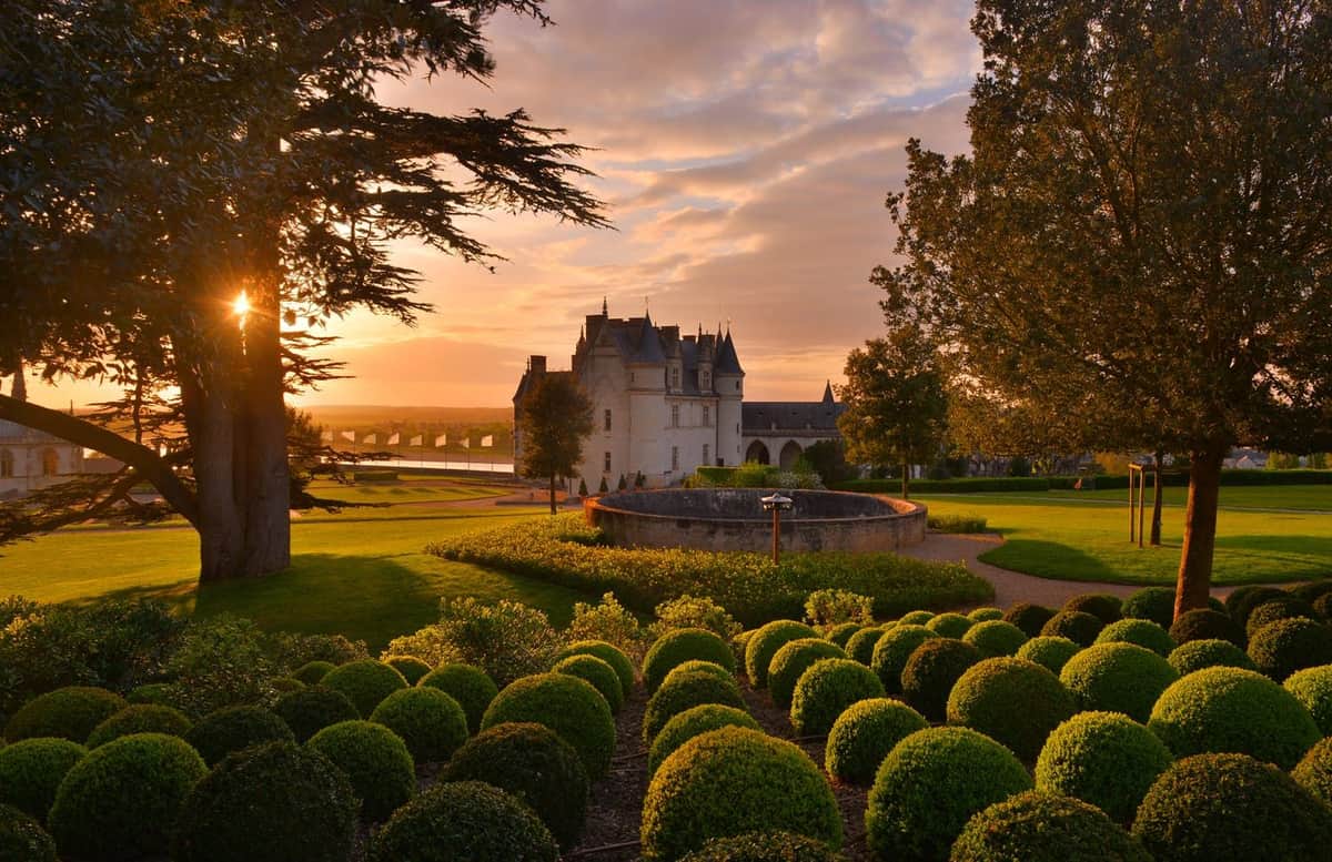 Manicured garden with round hedges, trees, and a stone building in the background at sunset.