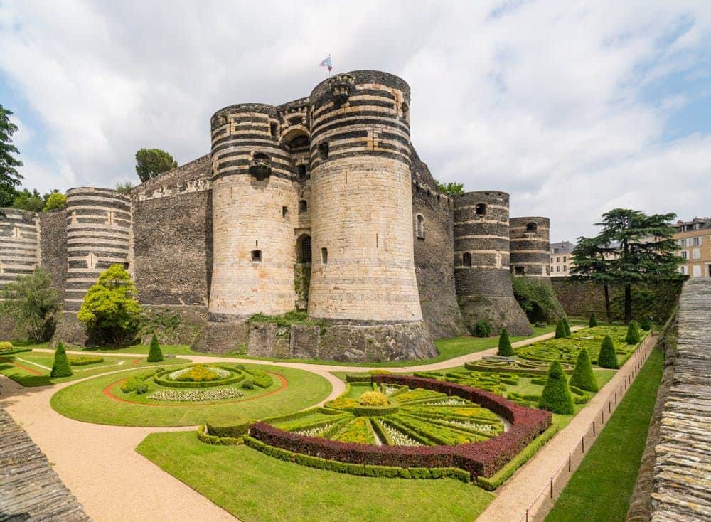 Medieval castle with striped stone towers and manicured gardens with geometric designs under a cloudy sky.