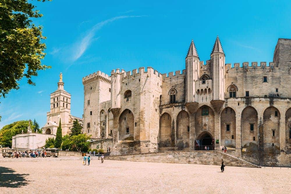 Palace of the Popes & Pont d’Avignon: Entry Ticket Historic castle with tall towers and crenelated walls under a clear blue sky, with a few people walking on the large courtyard below.