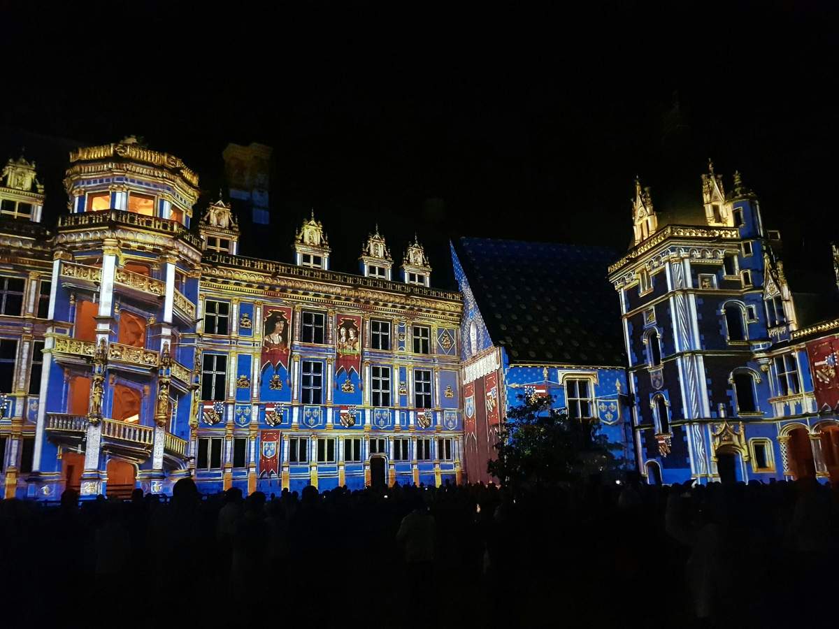 Royal Château de Blois: Sound and Light Show A building illuminated with vibrant blue, red, and gold lights against a dark night sky, with a crowd of people in front.