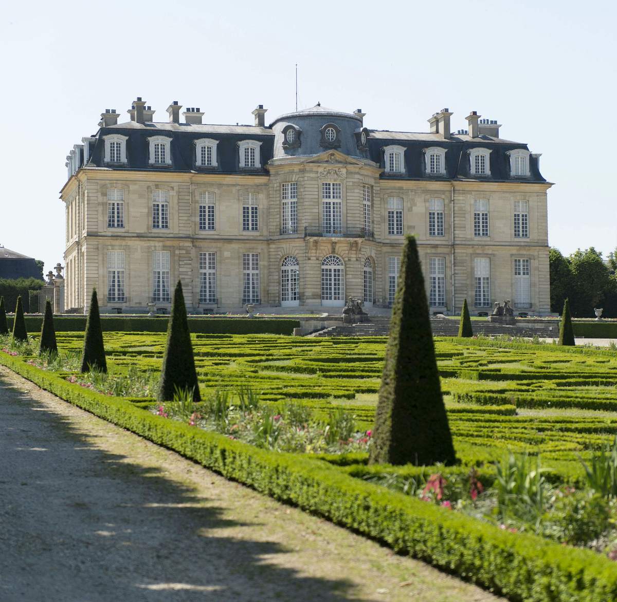 Formal garden with manicured hedges and topiary trees in front of a large, ornate mansion with multiple windows and a steep roof.