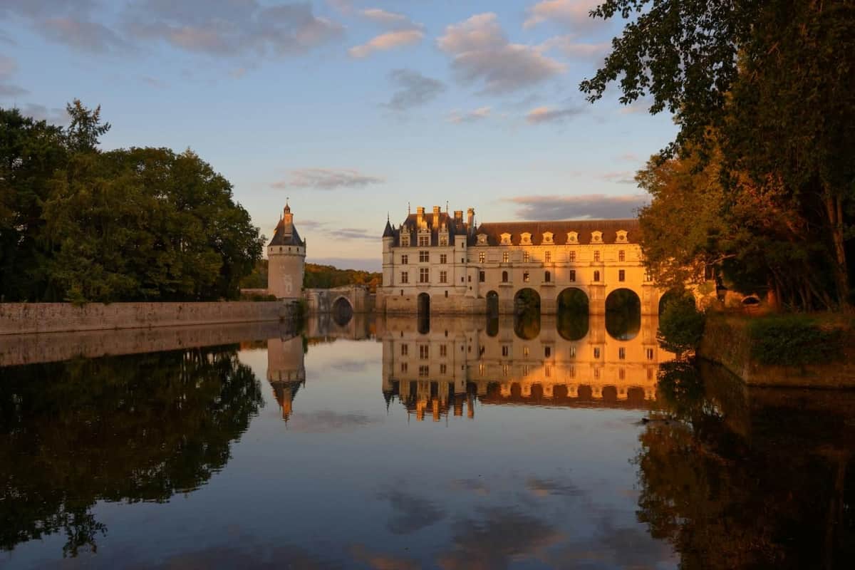 Château de Chenonceau: Entry Ticket Castle with arched bridges over a reflective river, trees on both sides, and a sunset-lit sky with clouds.