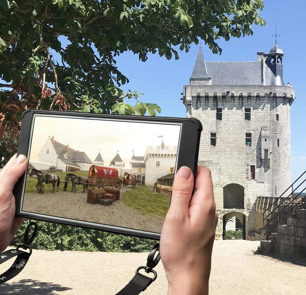 A person holds a tablet displaying a medieval scene in front of a real medieval castle with a clear blue sky.