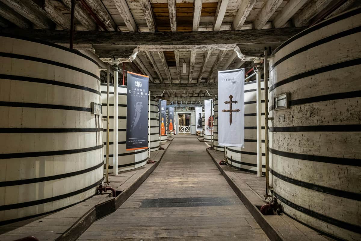 A wooden walkway flanked by large, striped barrels in a rustic building, with hanging banners and a beamed ceiling.