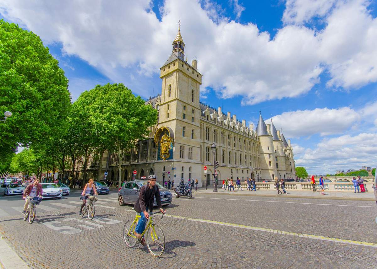 Conciergerie: Entry Ticket People biking and walking near a historic, ornate building with a clock tower and pointed turrets under a bright, partly cloudy sky.
