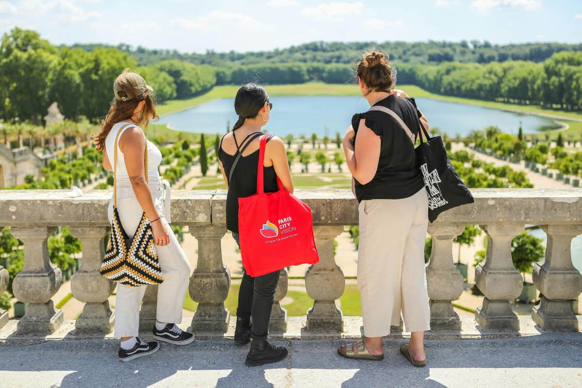Three women stand on a terrace overlooking a formal garden with a pond and forest in the distance.
