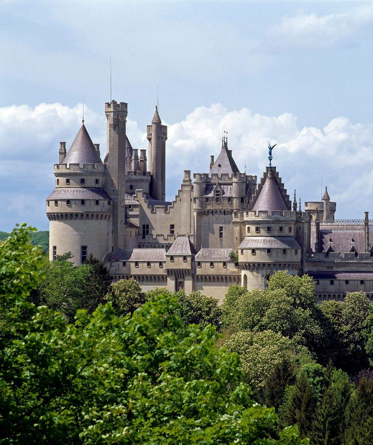 A medieval stone castle with multiple towers and turrets, surrounded by dense green trees under a partially cloudy sky.