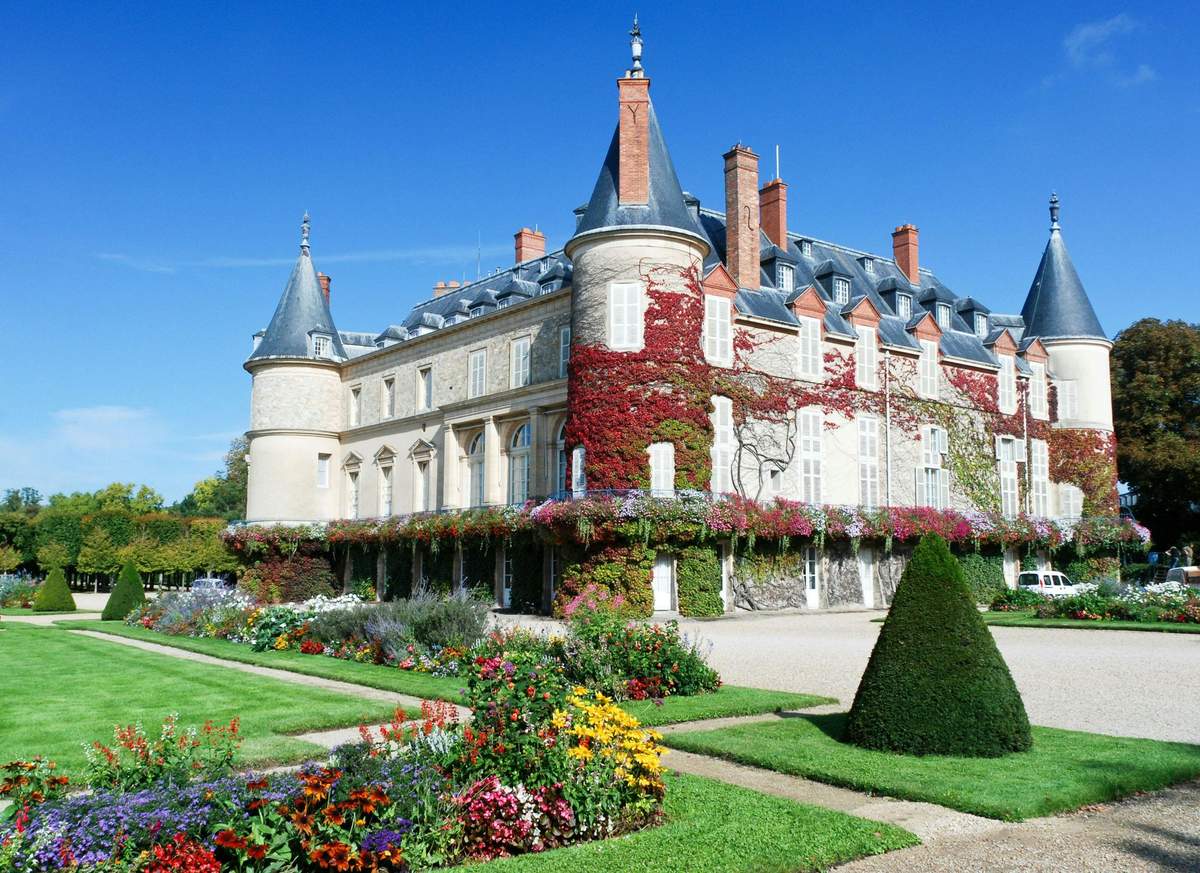 A historic building with ivy climbing its walls, surrounded by a manicured garden with colorful flowers and a trimmed shrub.