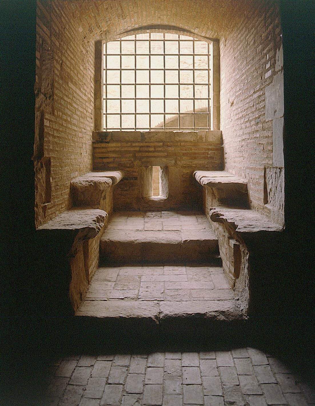 A narrow stone room with arched steps and two stone benches, lit by a small window with metal bars.