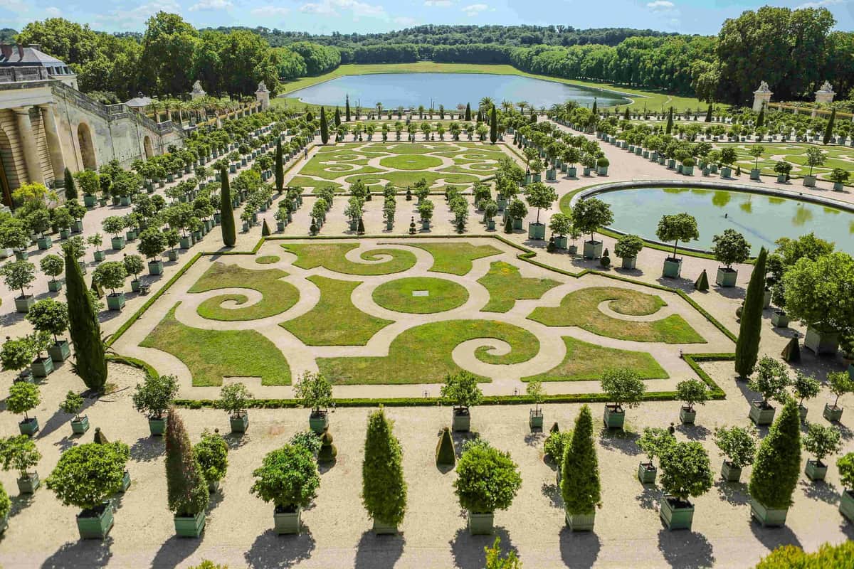 Ornate garden featuring symmetrical, geometric hedges, potted trees, and a central pond, with a forest and sky in the background.
