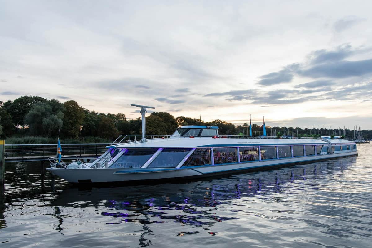 A modern glass-walled boat docked on calm water near a tree-lined shore at sunset, with reflections in the water.