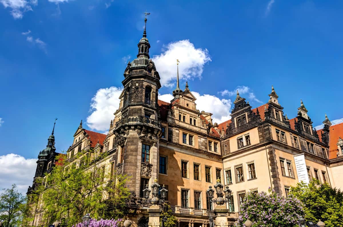 Historic building with ornate architecture, spires, and large windows under a blue sky with clouds. Trees and purple flowers in front.