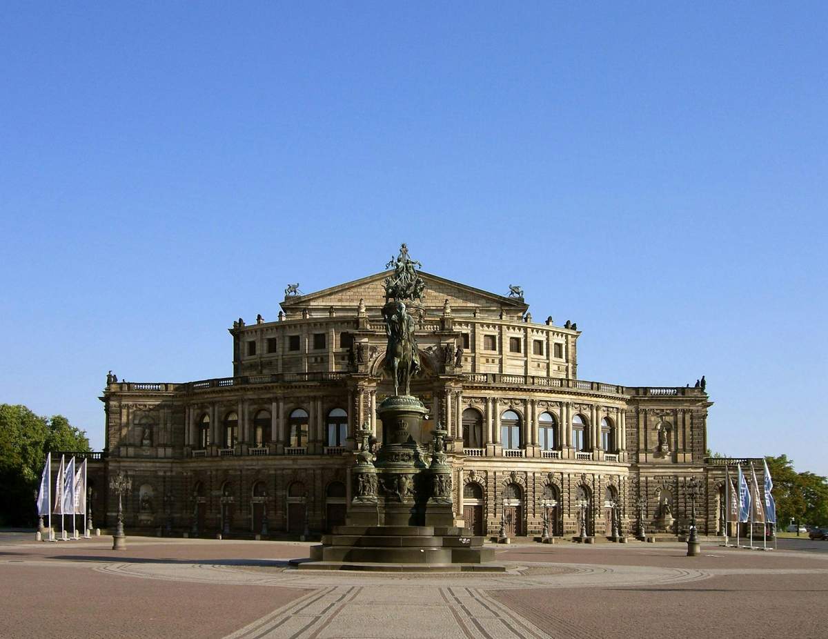 Historic building with ornate architecture, large arched windows, and a central statue set in a spacious plaza under a clear blue sky.