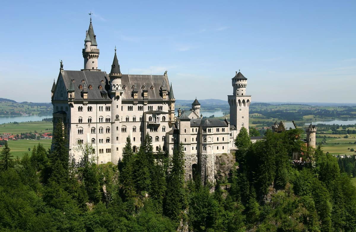 A large, ornate castle with multiple towers and turrets, surrounded by lush green trees, with a distant view of a lake and countryside.