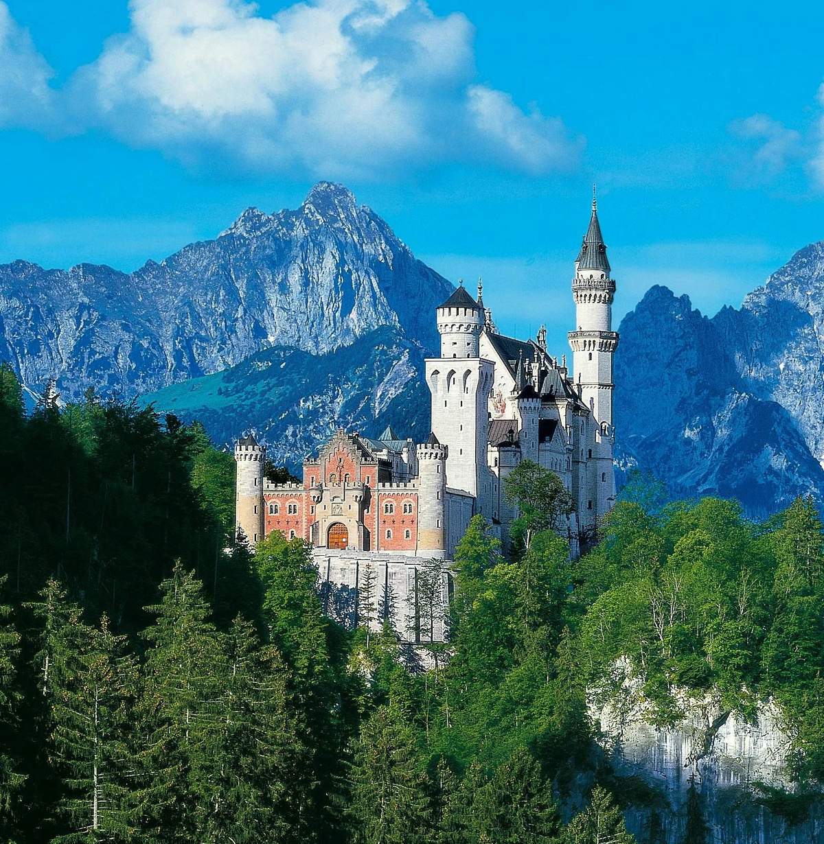 The Royal Castles Neuschwanstein & Linderhof: Day Trip from Munich Castle with multiple towers surrounded by dense forest, set against a backdrop of large, rocky mountains under a blue sky.
