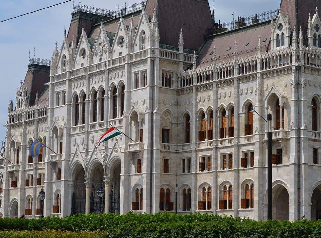 Budapest: Barrio del Castillo y Parlamento Húngaro Visita Guiada + Transporte Edificio blanco ornamentado de estilo gótico con ventanas arqueadas, detalles intrincados y una bandera, situado detrás de un seto verde.