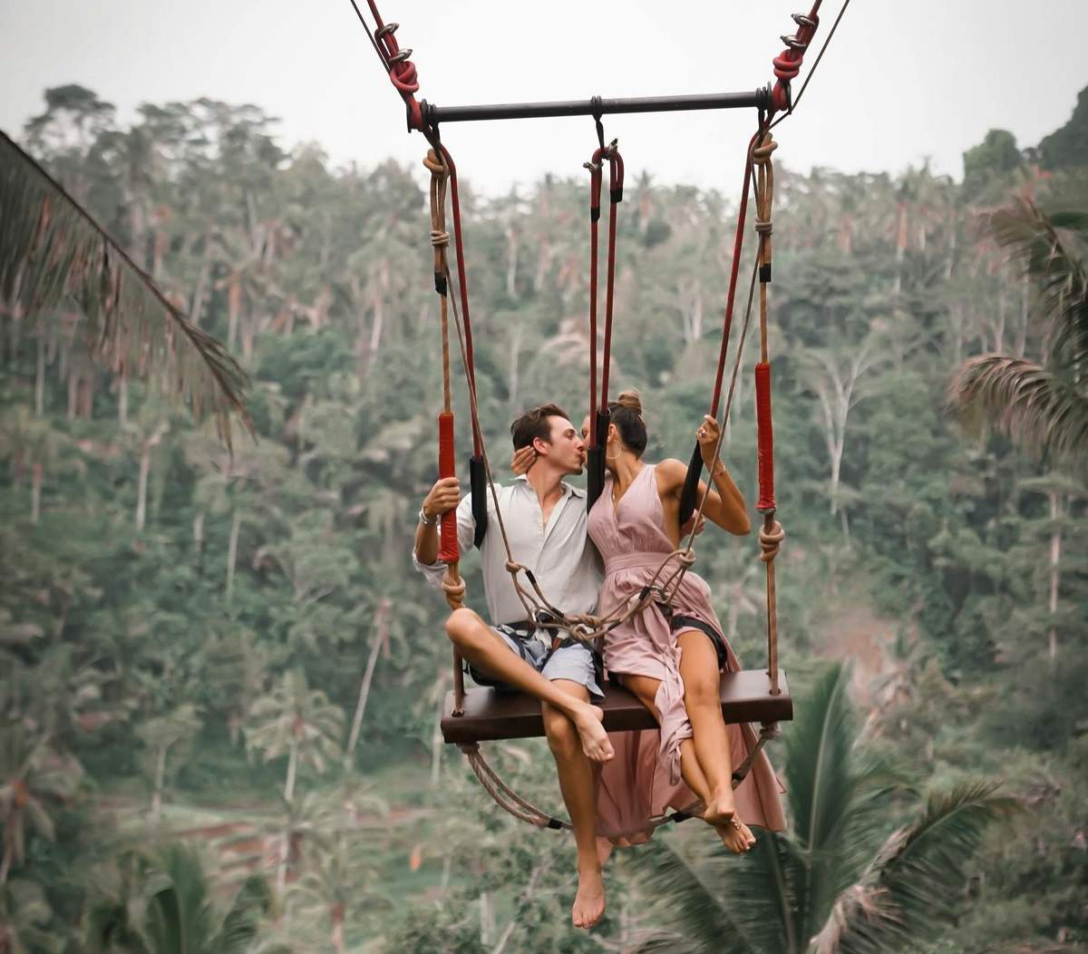 Best of Ubud: Monkey Forest, Rice Terraces, Tegenungan Waterfall & Temples A couple sits on a swing suspended high above a lush, tropical forest, sharing a kiss.