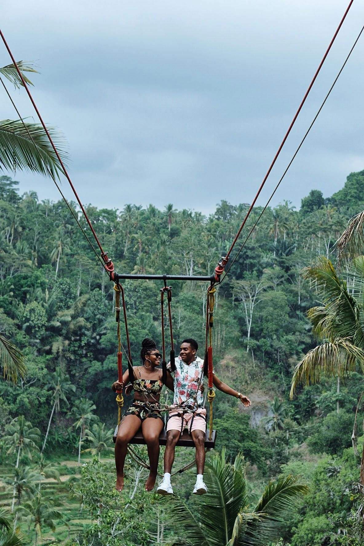 Two people sitting on a swing suspended high above lush green forest terrain with dense palm trees, smiling and holding hands.