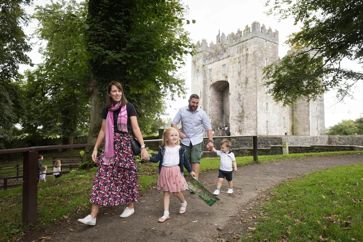 A family of four walks on a path near an old stone castle, surrounded by trees and greenery.