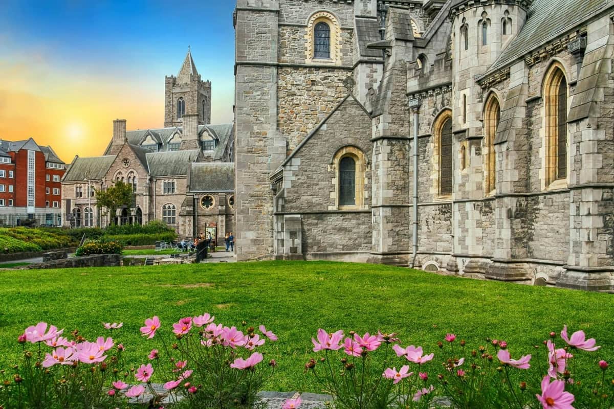 Libro de Kells, Castillo de Dublín y Catedral de Christ Church: Visita guiada a pie Histórica iglesia de piedra de arquitectura gótica, plantas rosas en flor en primer plano, césped bien cuidado y un cielo azul al atardecer.