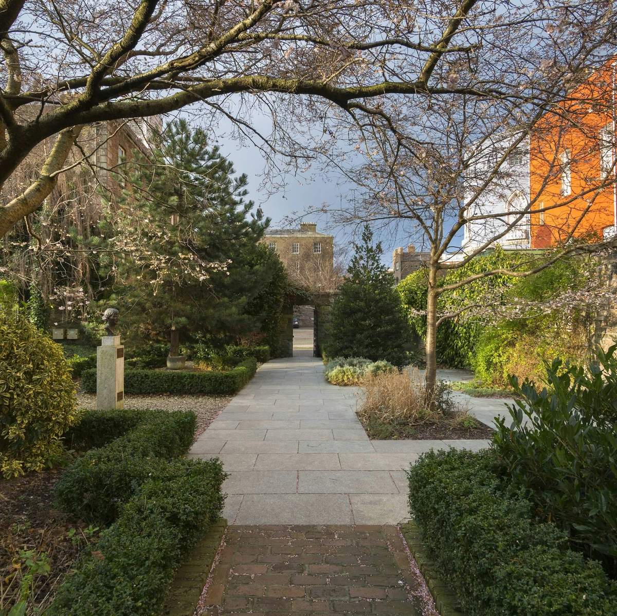 A garden pathway lined with hedges and trees, leading to a building in the background. Bust sculpture is visible on the left.