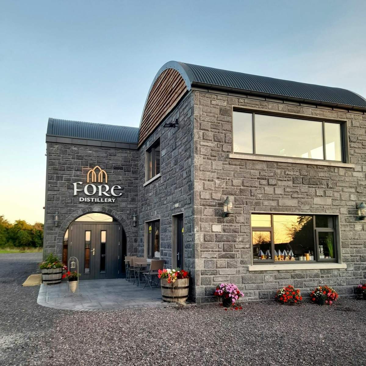 A stone building labeled "Fore Distillery" with large windows, outdoor seating, potted plants, and a gravel pathway.