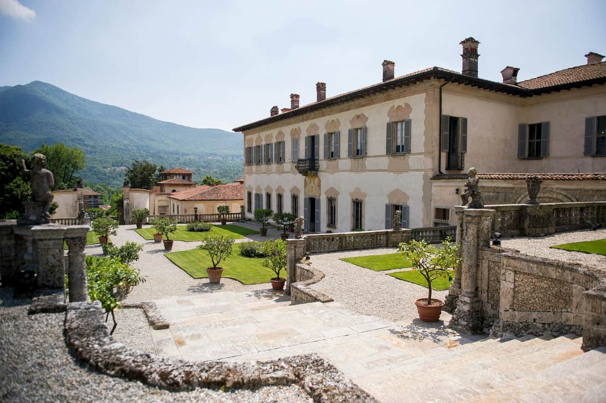 Villa Della Porta Bozzolo: Entry Ticket A historic villa with beige walls, green shutters, and manicured gardens set against a mountainous backdrop on a sunny day.