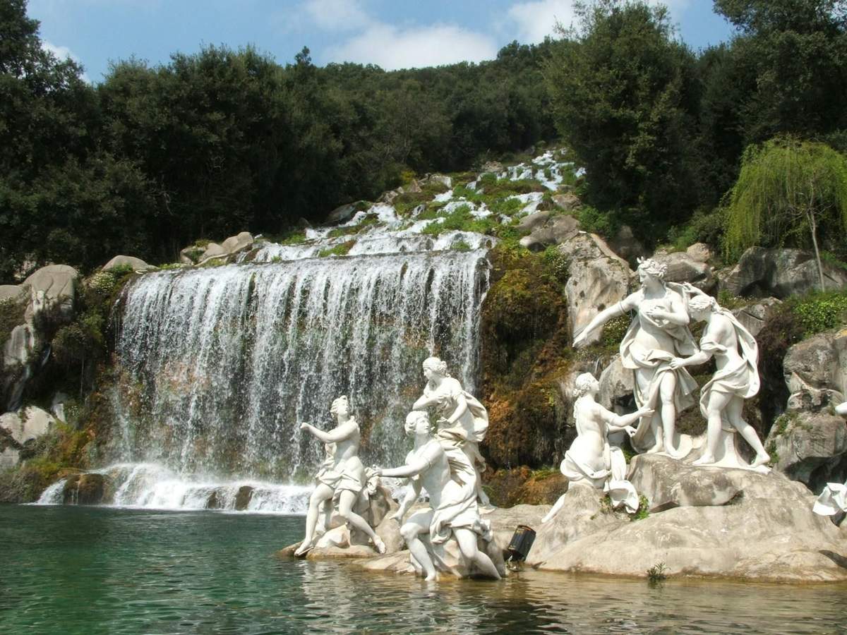 White statues depicting human figures stand near a cascading waterfall surrounded by lush greenery.